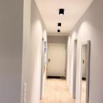 Flur mit hellen Wänden, Holzboden und Deckenleuchten. Türen zu verschiedenen Räumen sind sichtbar.