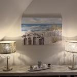 Wandbild mit Strandmotiv über einem Tisch mit zwei Lampen und Pflanzen.