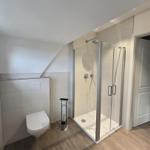 Dusche mit Glaswand, WC und Holzfußboden in einem hellen Badezimmer.