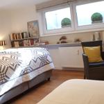 Schlafzimmer mit Bett, Sessel und Fenster mit Pflanzen.