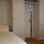 Zimmer mit zwei Betten, Kleiderschrank und Lampen. Tür führt in einen Flur.