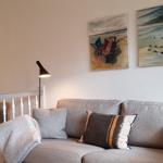 Grauer Sofa mit Kissen und Decke. Wandbilder zeigen Strand. Stehende Lampe und Holzregal.