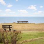 View through a window to a beach with two wooden benches and a blue sea under a clear sky.