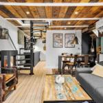 Wohnbereich mit Kamin, Sofa und Esstisch. Holzdecke und -boden. Treppenhaus im Hintergrund.