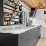 Küche mit Holzdecke, weißen Wänden und grauen Schränken. Aufbewahrungselemente und Küchengeräte sind sichtbar.