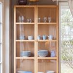 Wooden cabinet with glass doors and dishware. Windows and ceiling with wood paneling.