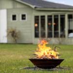 A burning fire pit on a lawn in front of a house with a porch.