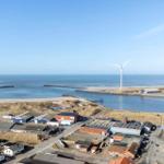 Meer, Windräder und Hafenbereich mit Industriegebäuden und Wohnhäusern.
