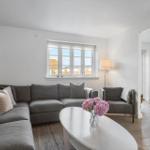 Wohnzimmer mit grauem Sofa, weißem Tisch und Blumen. Fenster zeigen Blick auf Gebäude.
