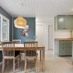 Dining area with wooden table, chairs, and kitchen. Windows with view of forest.
