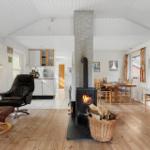 Living area with wood stove, dining table, and kitchen. Wooden floor and white walls.