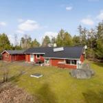 Red wooden house with dark roof, surrounded by grass and trees.