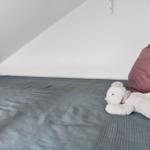 Ein Bett mit grauer Bettwäsche und einem weißen Plüschtier. Hintergrund ist weiß und schräg.
