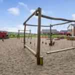 Großer Sandplatz mit Holzspielgeräten und Rutschbahn im Hintergrund.