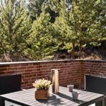 Terrasse mit Tisch, Stühlen, Thermoskanne und Pflanze. Hintergrund: Ziegelmauer und Nadelbäume.