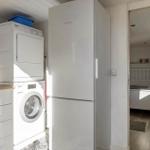 Kitchen with washing machine, dryer, and refrigerator. Corridor leading to bedroom with bed.