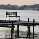 A bench sits on a wooden platform over water. Hills and trees are visible in the background.