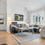 Living room with sofa, table, and chair. Windows show garden and house.
