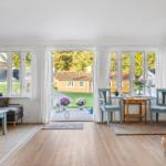 A room with wooden floor, windows, and a view of a garden. There is a table with two chairs and a guitar.