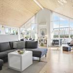 Living room with gray sofa, white table, and wooden ceiling. Large windows and light.