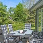 Terrace with table and chairs, surrounded by greenery and a house.