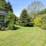 House with large garden and surrounding trees under blue sky.