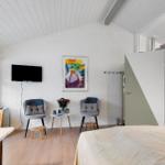 Room with bed, TV, two chairs, and window. Kitchen visible in background.