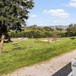 Green playground with slide and table under trees. View of water and forest.
