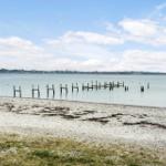 Beach with wooden posts in water and island in background under blue sky.