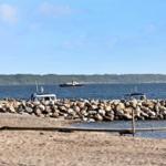 Beach with stone breakwater and boats. In the background, sea and forested shore.