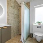 Badezimmer mit Dusche, Waschbecken und Toilette. Steinwand und Fenster sichtbar.