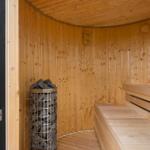Holzsauna mit Sitzbank und Steinbehälter. Tür offen mit Blick auf Grünfläche.