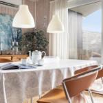 Dining table with tablecloth, cups, and teapot. Bright lamps and large windows.