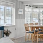 Dining room with wooden table, chairs, and windows. Walls with striped wallpaper.