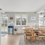 Dining area with wooden table, chairs, and striped wall. Windows and ceiling light visible.