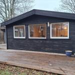 Schwarzes Holzhaus mit Holzterrasse und Fenstern. Im Inneren sind Möbel und Beleuchtung sichtbar.