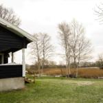 Schwarze Holzhütte mit Veranda, Grasfläche und Bäumen im Hintergrund.