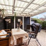Terrasse mit Tisch, Stühlen und Grill unter Glasdach. Wand mit Dekoration und Pflanzen.