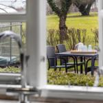 Durch ein Fenster blickt man auf eine Terrasse mit Tisch, Stühlen und einer Teekanne.