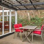 Terrasse mit Tisch und roten Sitzkissen unter Holzüberdachung. Blick in das Haus mit Essbereich.