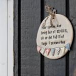 Ein Keramik-Schild mit Dänischer Aufschrift hängt an einer dunklen Holztür neben einem Fenster.