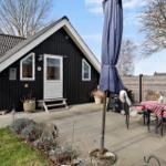 Schwarzes Holzhaus mit Terrasse und Garten. Tisch mit rot-weißem Tischtuch und Stühlen.