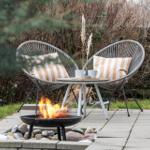 Zwei Sessel mit Kissen und Tisch stehen neben einem brennenden Feuerkessel im Garten.