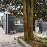 Ein graues Holzhaus mit weißer Tür und Fenstern. Außenbereich mit Sitzgelegenheit und Bäumen.