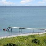 Ein Steg führt ins Meer. Eine Person taucht im Wasser. Windkraftanlagen sind im Hintergrund sichtbar.