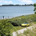 Ein gelbes Auto steht an einem Strand mit Gras und Blumen. Zwei Personen gehen am Wasser entlang.