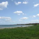 Strand mit Grasfläche und Häusern am Horizont unter blauem Himmel mit weißen Wolken.