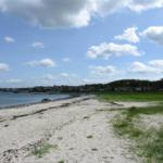Strand mit Sand, Gras und Häusern am Wasser unter blauem Himmel mit Wolken.
