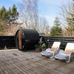 Deck with sauna, lounge chairs, and wooden railing. Background: trees and clear sky.