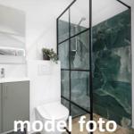 Badezimmer mit Dusche, Toilette und Waschbecken. Grüne Marmorwand und Fenster mit Pflanze.